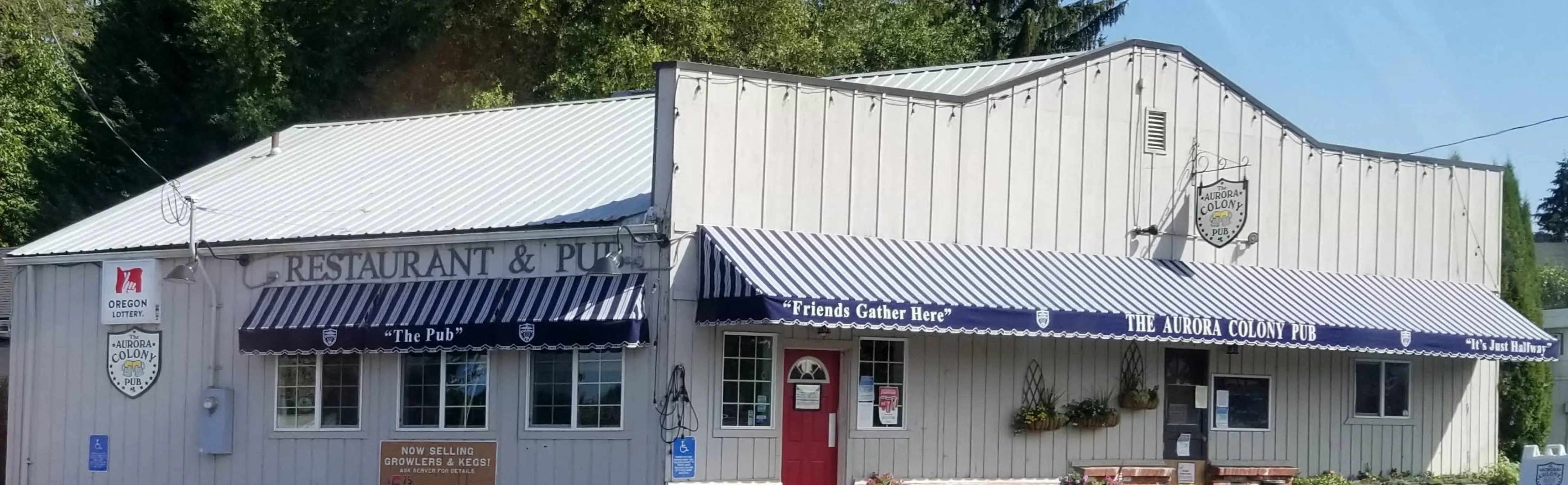The Aurora Colony Pub in daylight — a long white building with blue-striped awnings and 'Friends Gather Here' over the entrance.