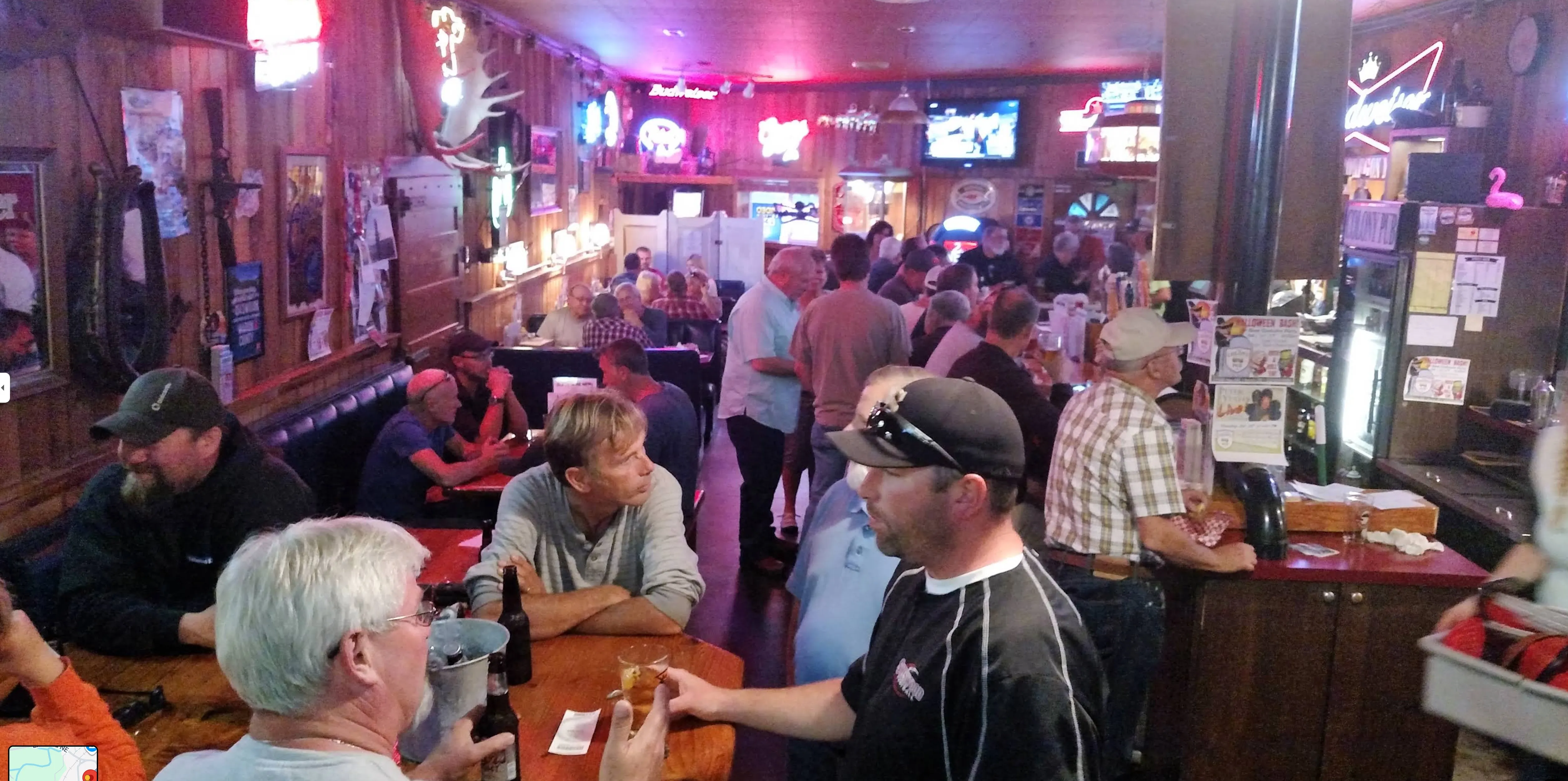 A packed crowd fills the room — friends laughing over beers beneath glowing neon, a mounted deer head and warm wood paneling.