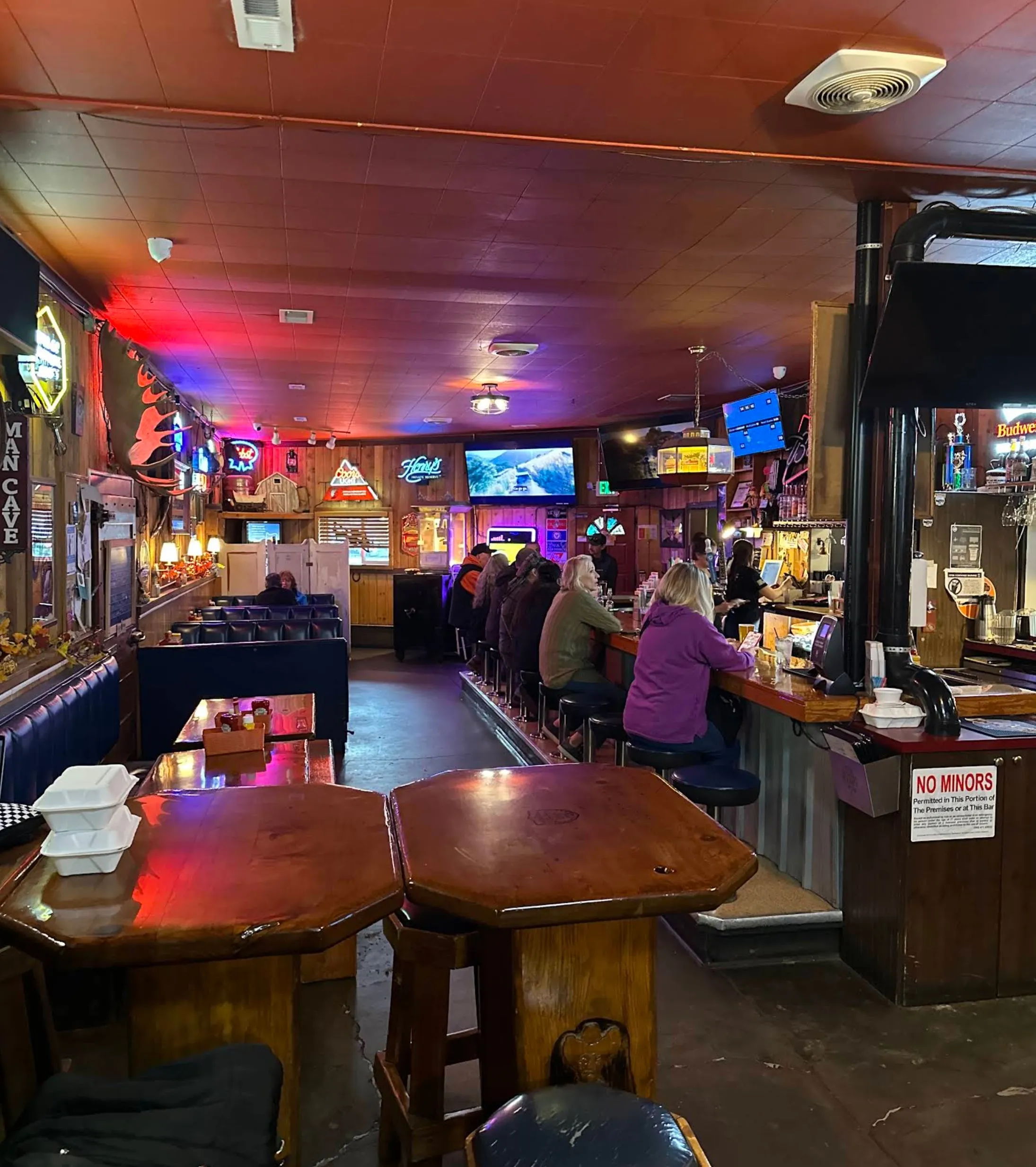 The long bar seen end to end — patrons on stools, neon signs down the wall and the red ceiling glow over polished wood tables.